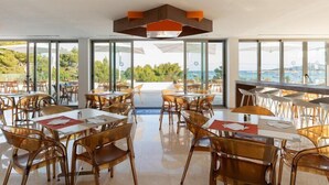 A restaurant with tables and chairs, large windows, and a view of the sea.