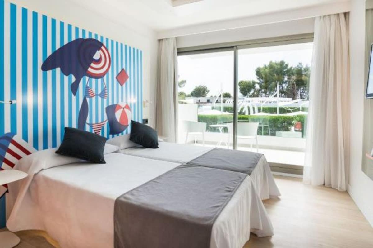 A hotel room with a large bed, two pillows, a striped wall mural, and a balcony with outdoor furniture.