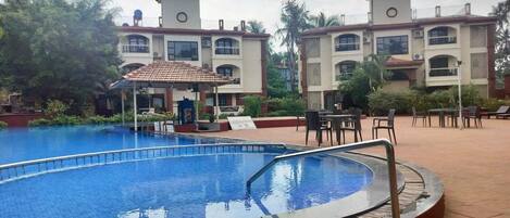 Una piscina con borde de baldosas, rodeada de mobiliario exterior y un edificio con varios balcones.