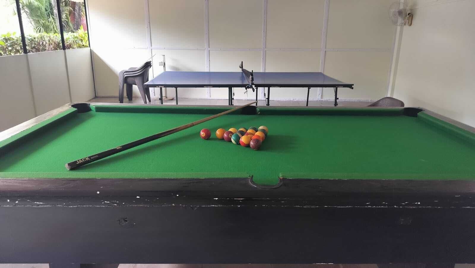 Een pooltafel met een keu en biljartballen, een tafeltennistafel, en een stoel in een ruimte met een witte muur en een raam.