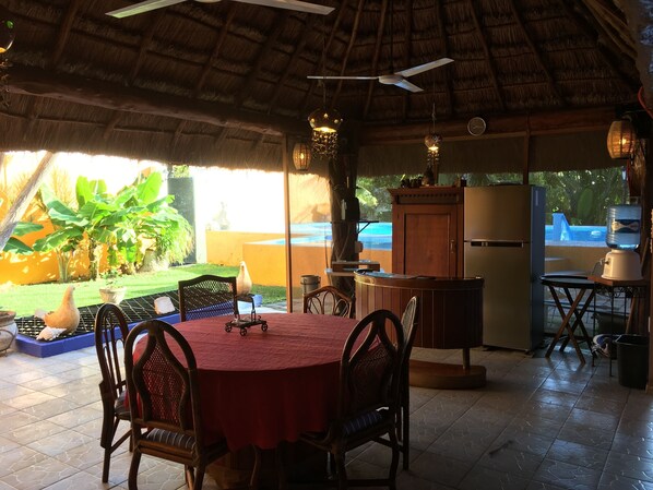 A covered outdoor dining area with a round table and chairs, a ceiling fan, and a view of a swimming pool.