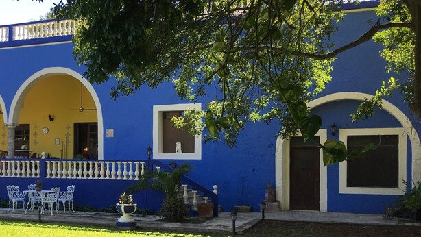 A building with blue and yellow exterior walls, white balconies, and a small garden area with a bench and palm trees. - Hacienda San Pedro Nohpat (Kanasín)