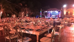 An outdoor dining area with multiple tables set for guests, surrounded by palm trees and a stage with colorful lighting in the background. - Hacienda San Pedro Nohpat (Kanasín)