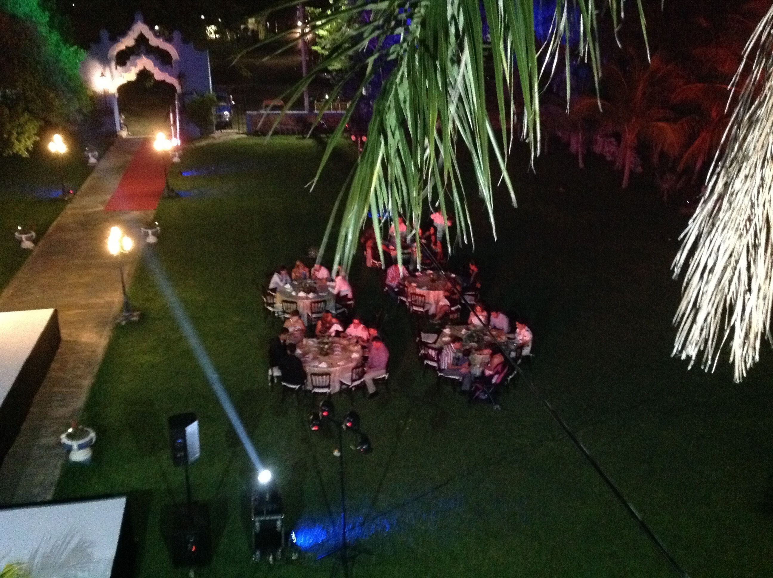 An outdoor dining area with tables and chairs arranged on a grassy lawn, illuminated by overhead lights.