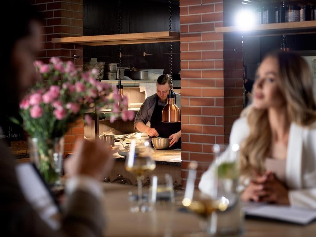 Ein Restaurantinnenraum mit Backsteinwand, einem Koch, der zubereitet, und Gästen an einem Tisch mit Weingläsern.