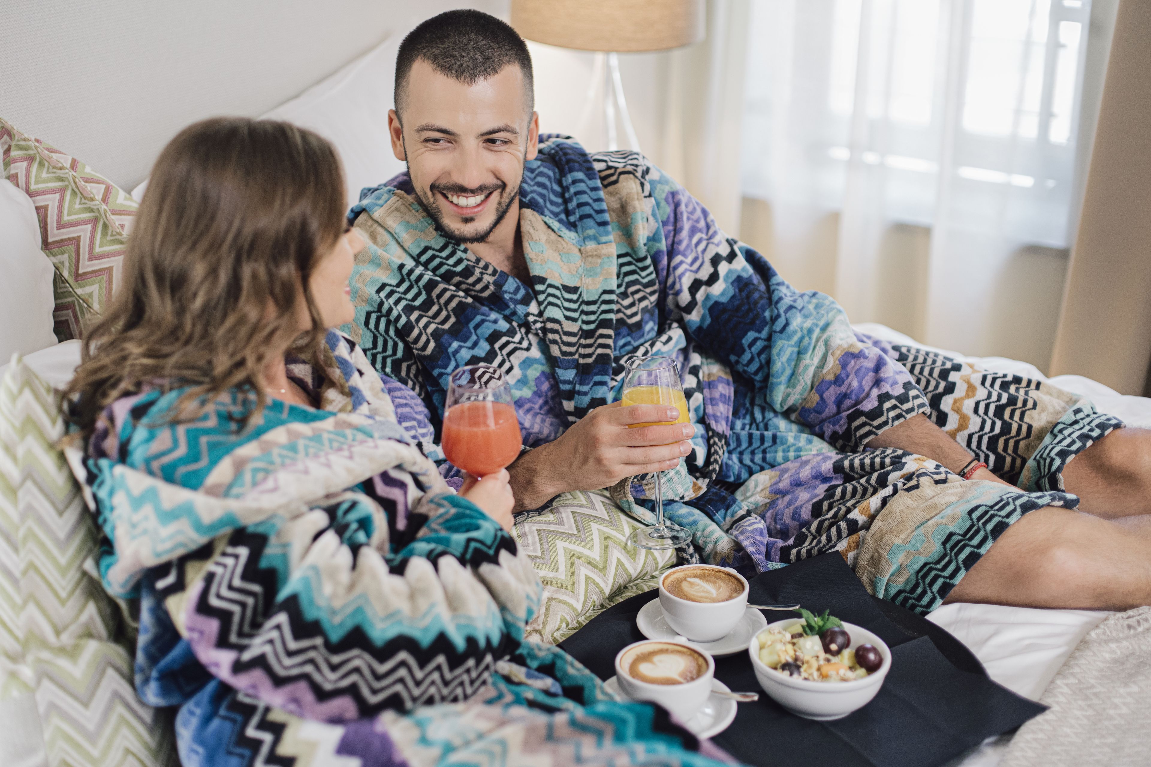 A couple in bed with a coffee table, a glass of orange juice, and a bowl of cereal.