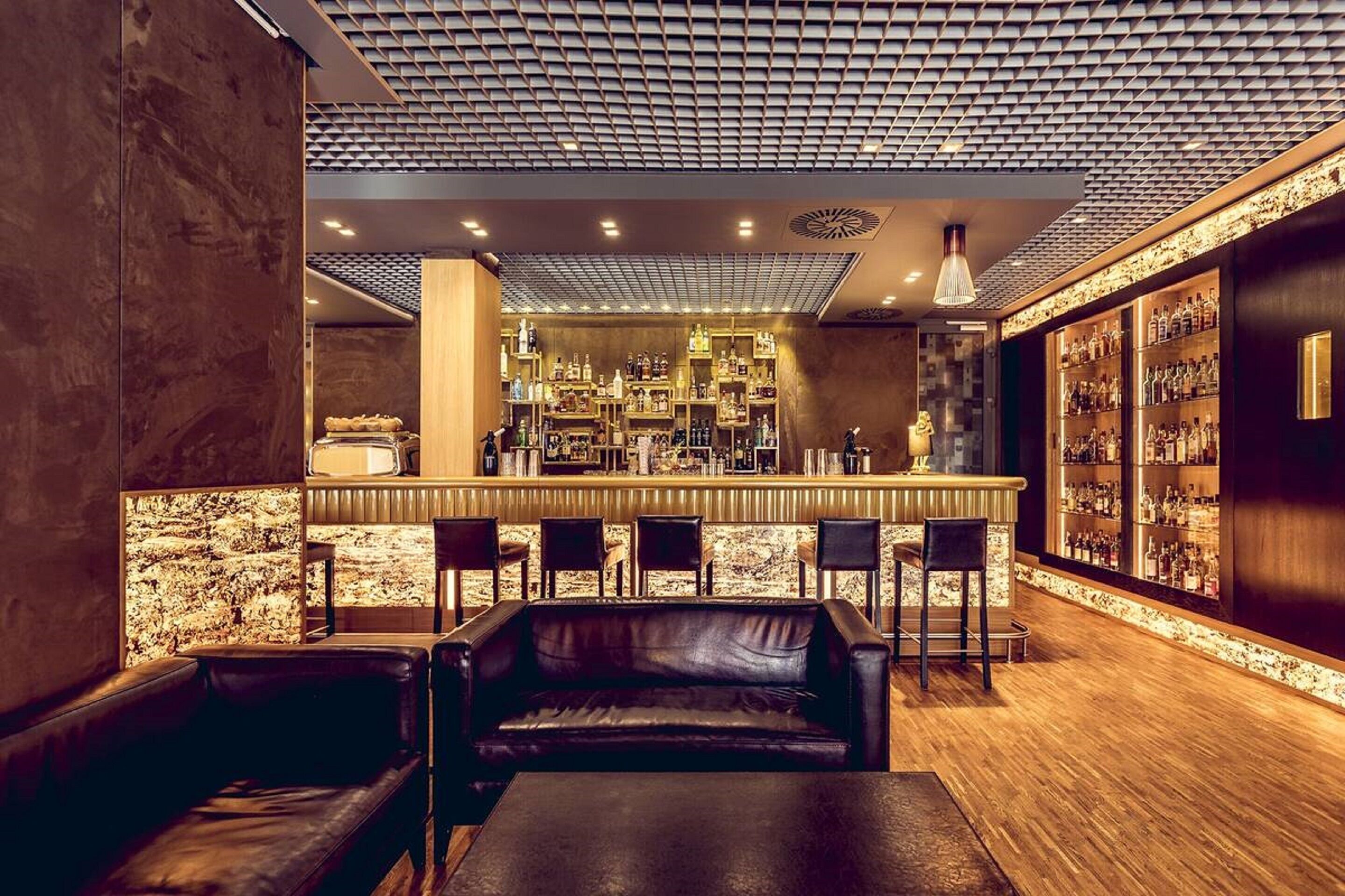 Uma área de bar com balcão de madeira, diversas opções de assento incluindo um sofá e cadeiras, e prateleiras repletas de garrafas.