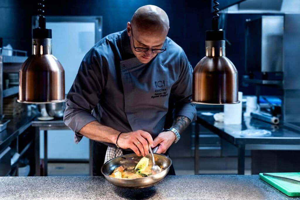 A chef in a professional kitchen with stainless steel appliances and pendant lighting.