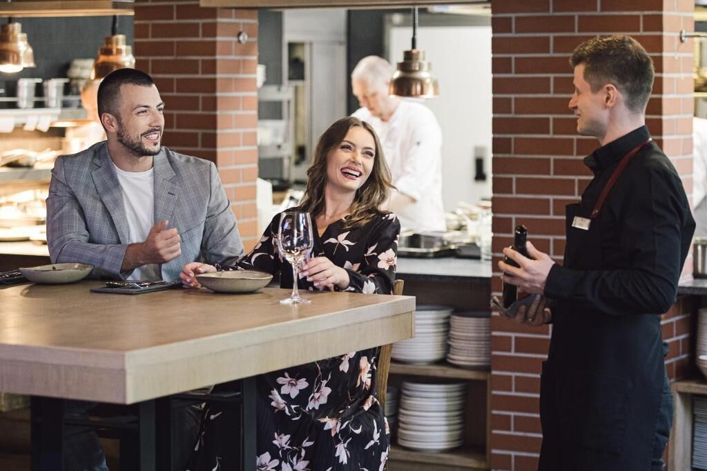 Ein Restaurantambiente mit Barbereich, ein Koch im Hintergrund und ein Kellner bedient einen Gast.