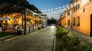 Un café en plein air avec des tables et des chaises sous des guirlandes lumineuses, un chemin pavé et des bâtiments de chaque côté.
