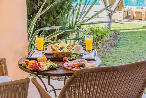 A breakfast table with fruits, pastries, cheese, and juice, set outdoors with a pool in the background.