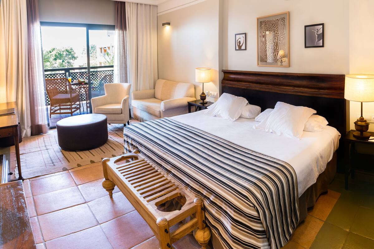 Ein Hotelzimmer mit einem großen Bett, einer Holzbank, einem Stuhl, einem Tisch und einem Balkon mit Blick auf Bäume.
