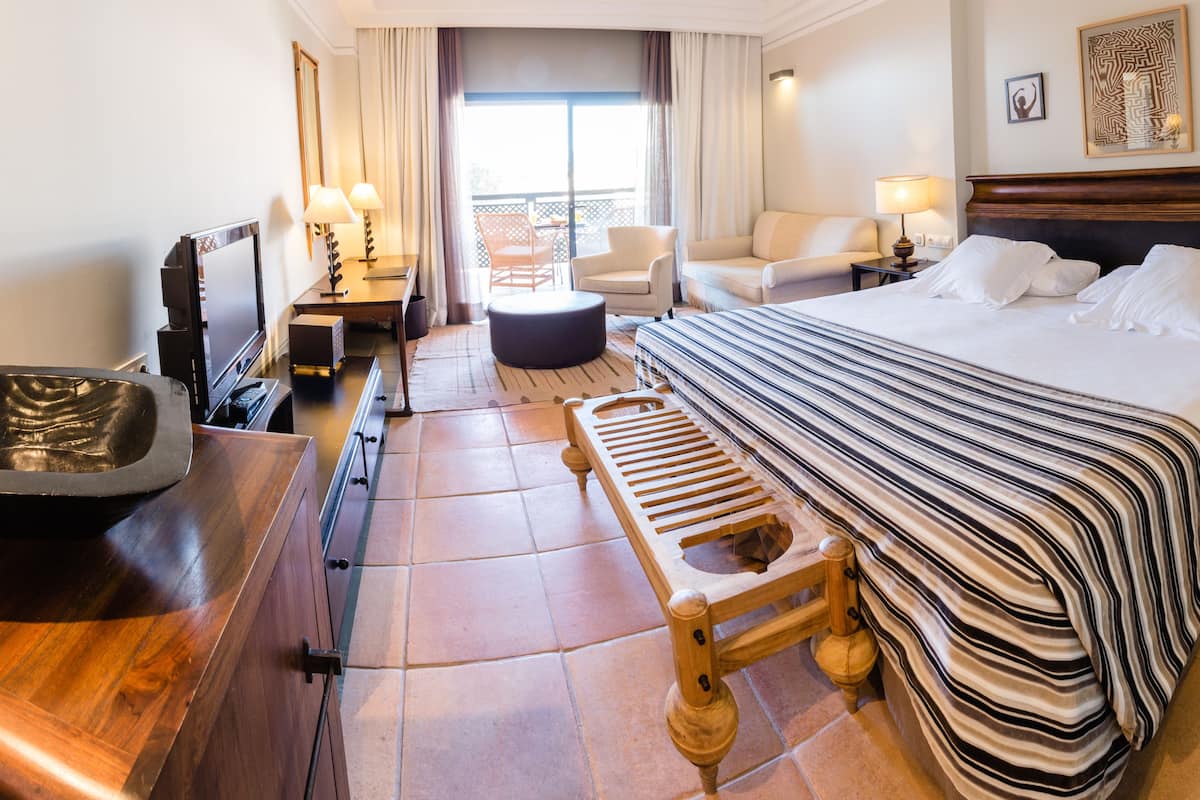 Ein Hotelzimmer mit einem großen Bett, einer hölzernen Bank, einem Schreibtisch mit Lampe, einem Sessel und Blick ins Freie.