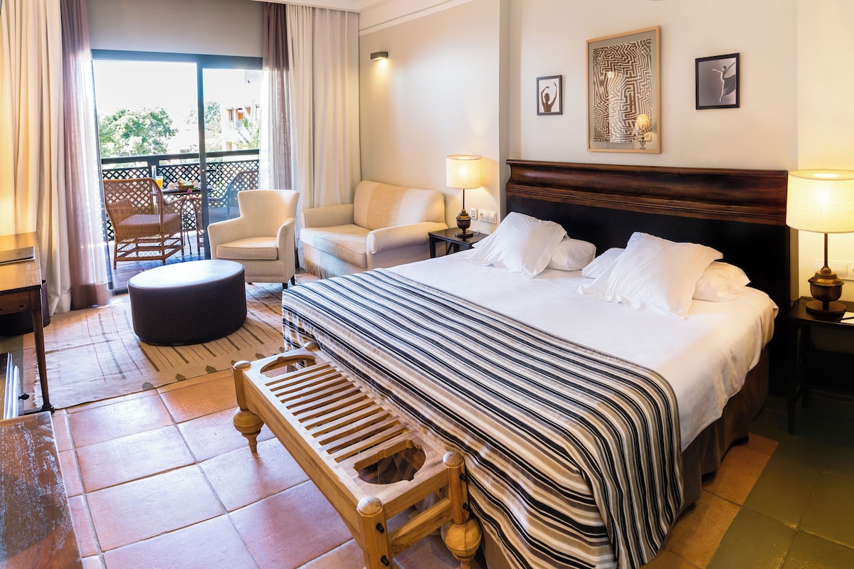 A hotel room with a large bed, a wooden bench, a chair, a small table, and a balcony with a view.