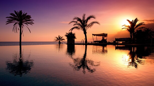 A serene sunset over a calm infinity pool with palm trees and a lounge area.