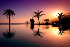 A serene sunset over a calm infinity pool with palm trees and a lounge area.