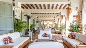 A spacious outdoor seating area with white sofas and a wooden coffee table, surrounded by potted plants and featuring a brick floor and wooden beams.