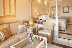 Habitación de hotel con sofá, una mesita con un florero, una silla de mimbre, cama con ropa de cama blanca y un espejo grande.