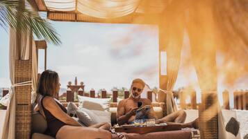 Una pareja descansando en una silla de mimbre con una bandeja de comida y bebidas, bajo una estructura cubierta con tela.