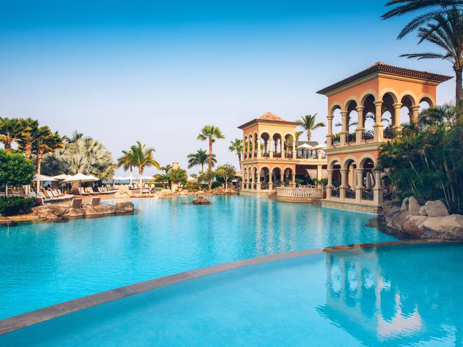 Una piscina grande con un edificio que tiene balcones arqueados y un techo de tejas.