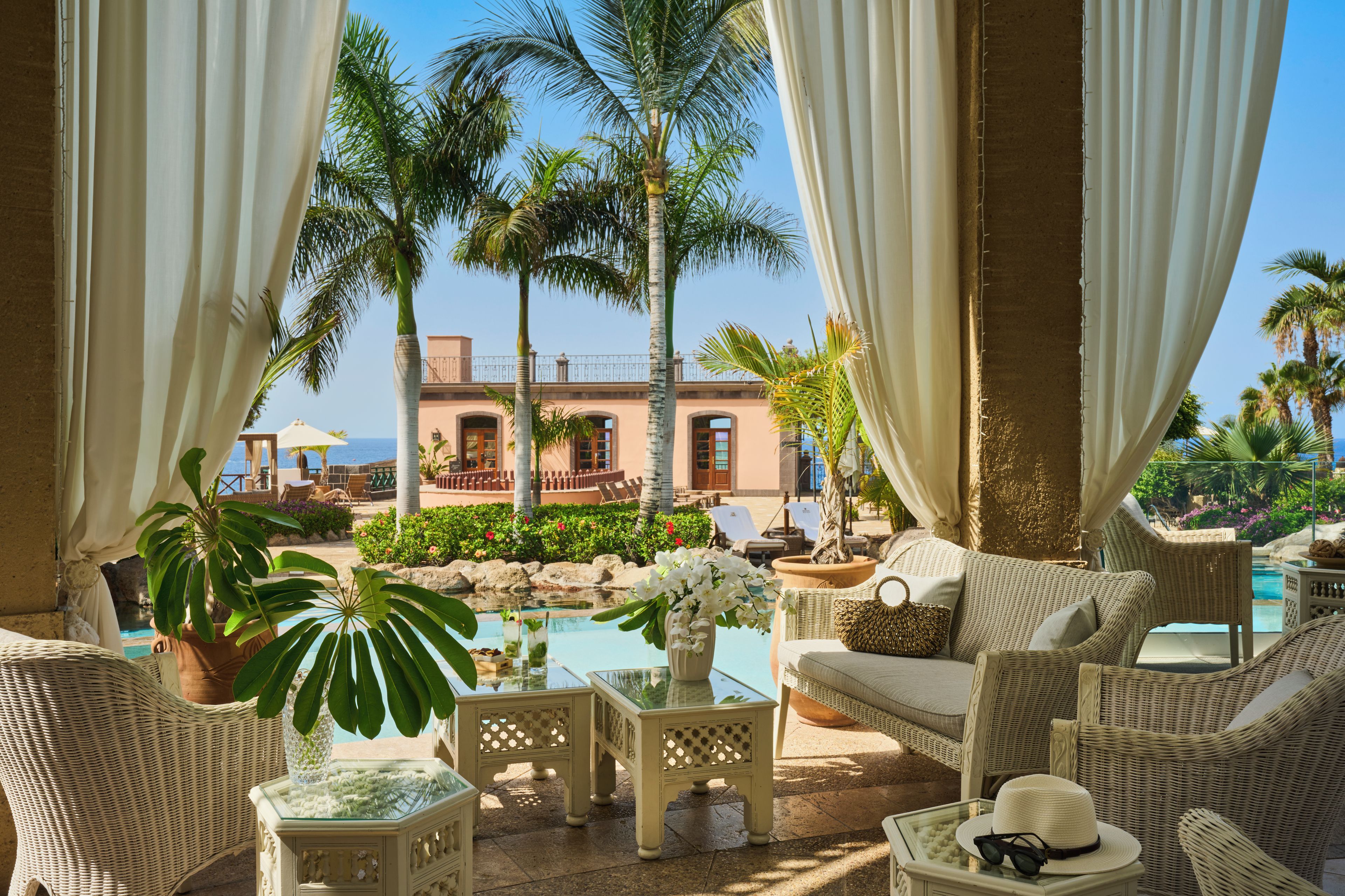 A poolside lounge area with wicker furniture, a glass-top table, and a view of a building and palm trees.