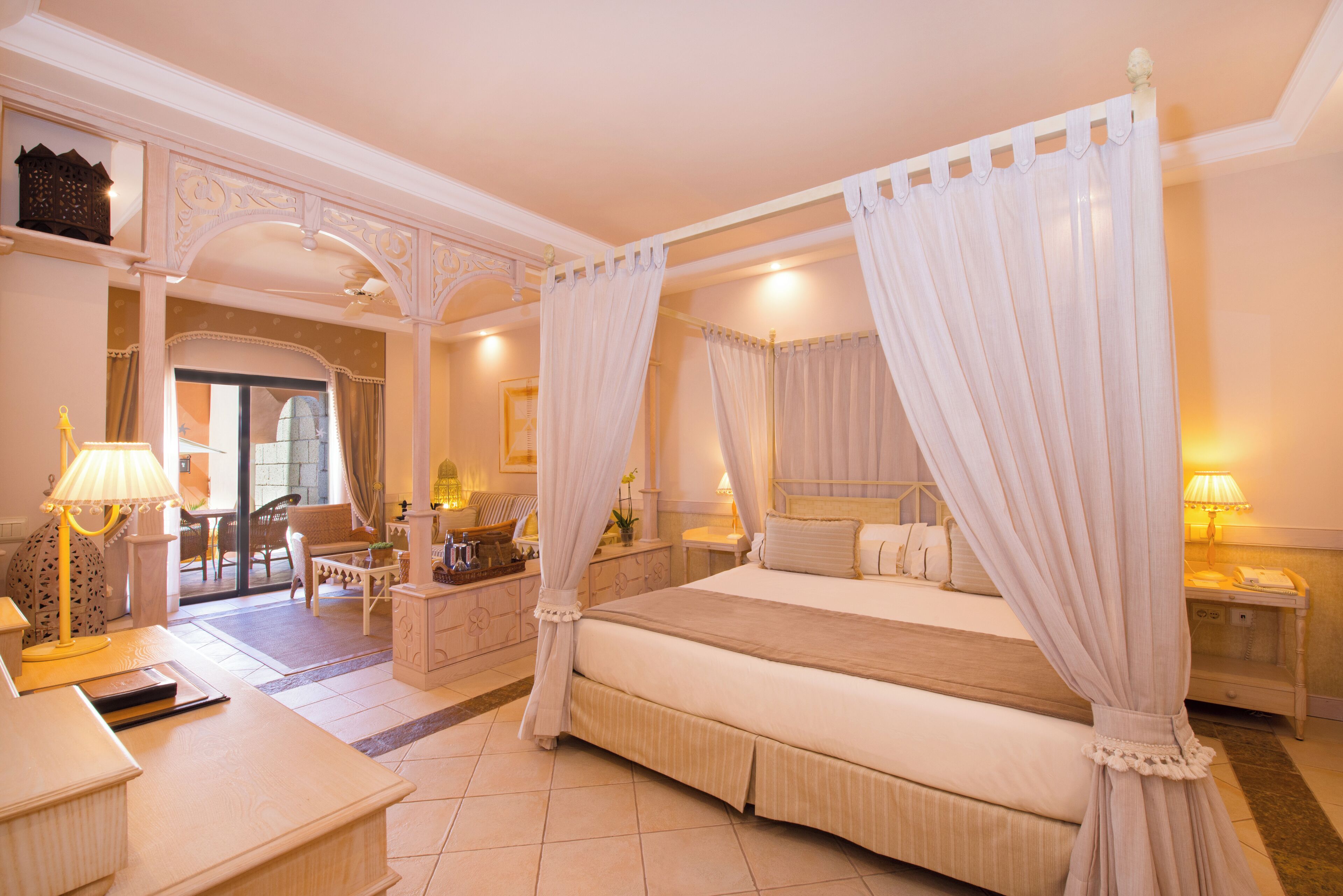 A spacious bedroom with a four-poster canopy bed, a sitting area with a sofa and armchairs, and a view of the outdoor patio.