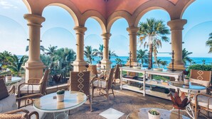 Un espace extérieur aménagé, abrité et doté de tables et de chaises, offrant une vue sur une plage bordée de palmiers et sur l’océan.