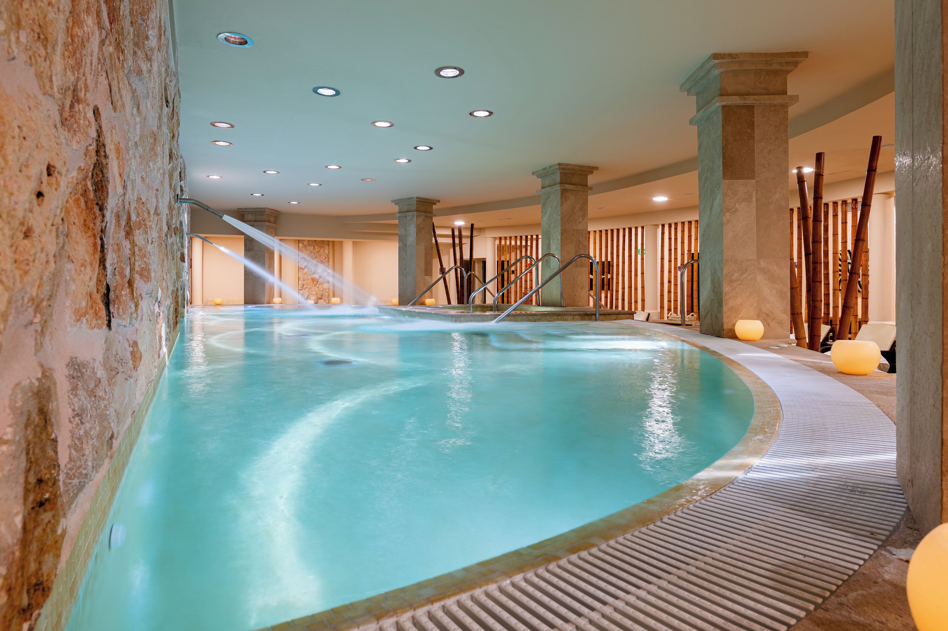 Una piscina interior con borde curvo, rodeada de muros y columnas de piedra, con un elemento acuático y una pared de bambú al fondo.