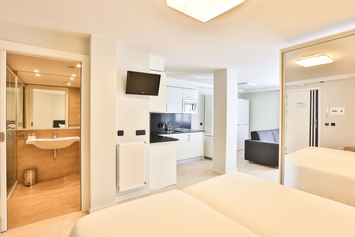 A modern hotel room with a large bed, a separate bathroom, a kitchenette, and a TV.