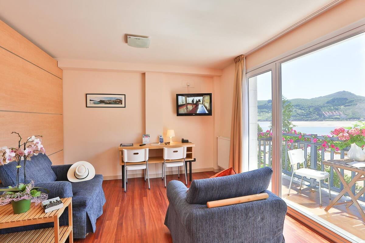 A room with a wooden desk, two chairs, a sofa, a TV, and a balcony with flowers.