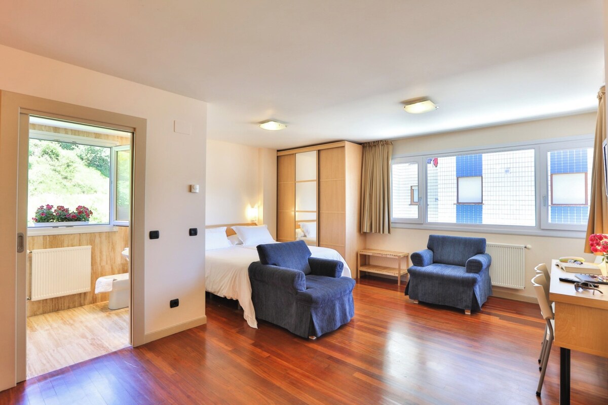 A modern hotel room with a bed, two armchairs, a desk, and large windows.