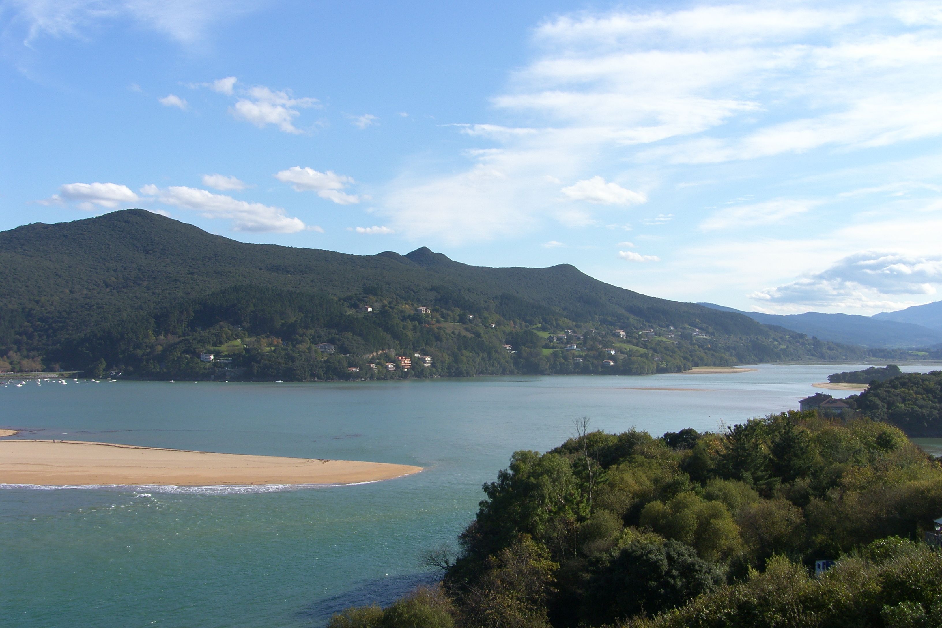 A coastal landscape with a beach, forested hills, and a body of water.