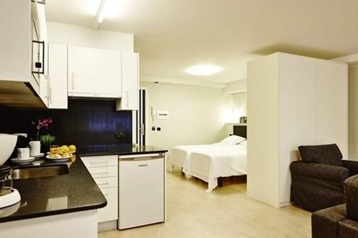 A compact hotel room with a kitchenette, a single bed, a small sitting area, and a separate bedroom.