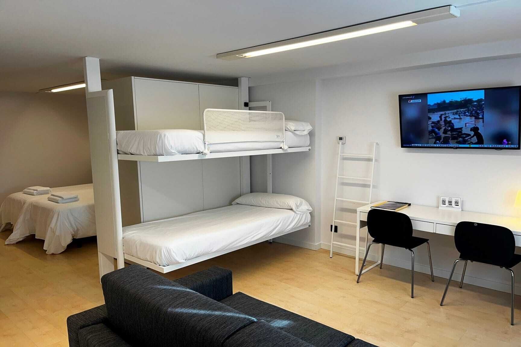 Habitación con literas, un escritorio, sillas, un televisor y una escalera.