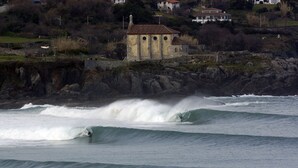 A coastal scene with a building on a rocky outcrop, waves crashing, and a surfer riding a wave. - Apartamentos Mundaka (Mundaka)