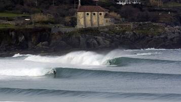 Un paysage côtier avec un bâtiment perché sur un promontoire rocheux, des vagues qui déferlent et un surfeur qui chevauche une vague.