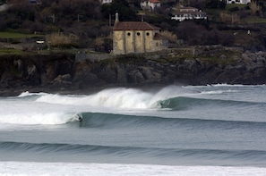 A coastal scene with a building on a rocky outcrop, waves crashing, and a surfer riding a wave. - Apartamentos Mundaka (Mundaka)