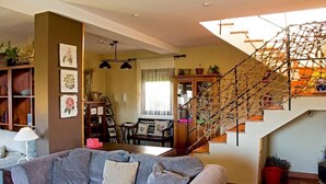 Una sala de estar con un sofá gris, una estantería de madera, una escalera con barandilla de madera y un comedor con mesa y sillas.