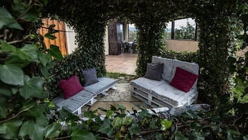 Zona d'estar a l'aire lliure amb dues plataformes blanques i seients mullidons, cadascuna amb un coixí vermell, envoltades de vegetació abundant.
