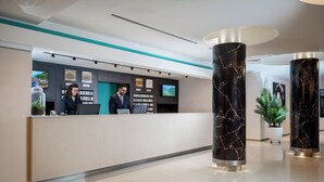 Een moderne hotel receptie met een balie, twee medewerkers, een groot digitaal scherm en decoratieve zuilen.