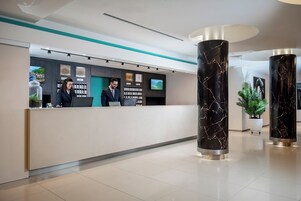 A modern hotel reception area with a reception desk, two staff members, a large digital screen, and decorative columns.