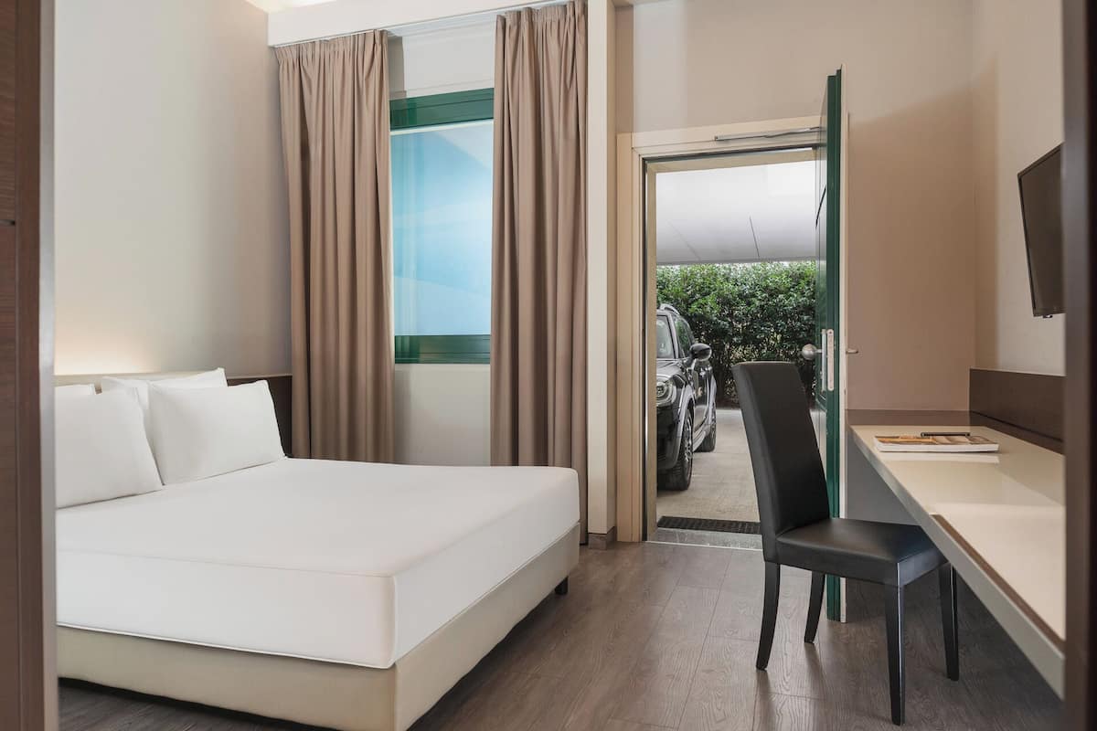 A hotel room with a bed, a desk, a chair, a TV, and a view of a car and greenery outside.