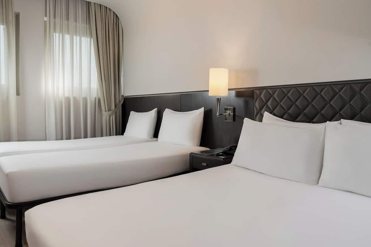 A hotel room with two beds, a nightstand with a lamp, and a window with sheer curtains.