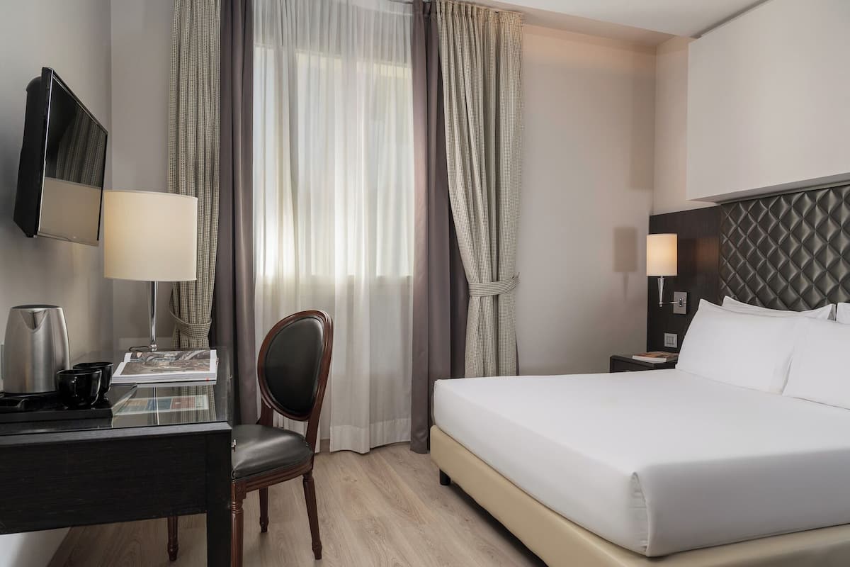 A hotel room with a bed, a desk with a lamp, a chair, a television, and curtains.