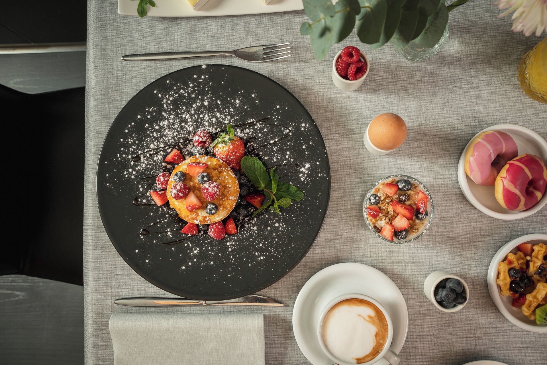 A breakfast setting with pancakes topped with berries and powdered sugar, accompanied by coffee and various pastries.