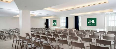 Una sala conferenze con file di sedie e tavoli, pareti bianche e luci a soffitto.
