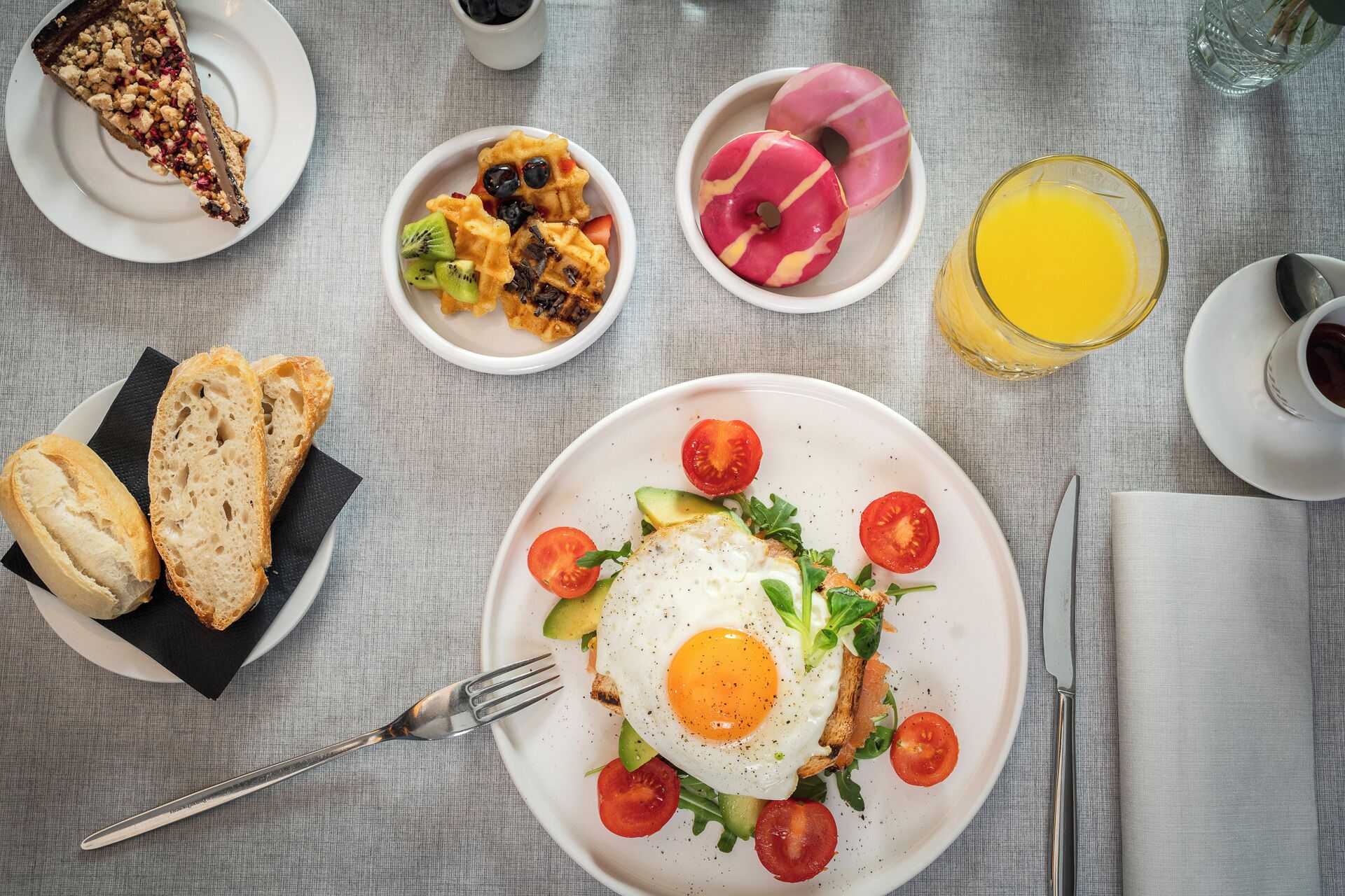 Un desayuno con huevos, tostadas y fruta, una rebanada de pastel, gofres, donas y un vaso de jugo de naranja.