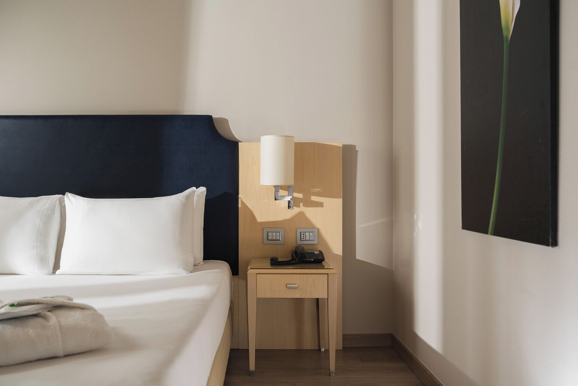 A hotel room with a bed, a bedside table with a lamp, a phone, and a small drawer.