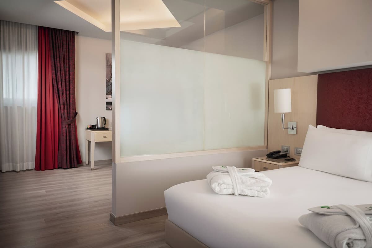 A hotel room with a bed, a nightstand, a desk, a chair, a red curtain, and a window with sheer curtains.