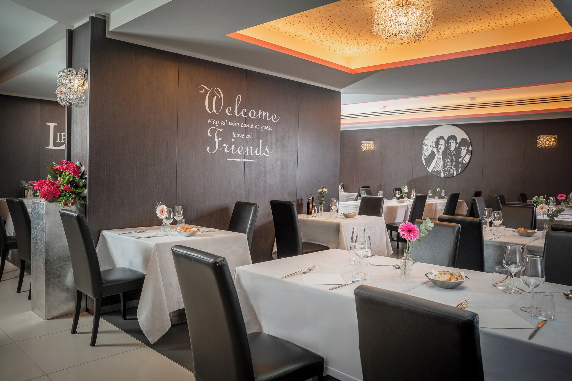 Een restaurant met meerdere eettafels, een bord met de tekst 'Welcome as Friends' aan de muur, en een kroonluchter.